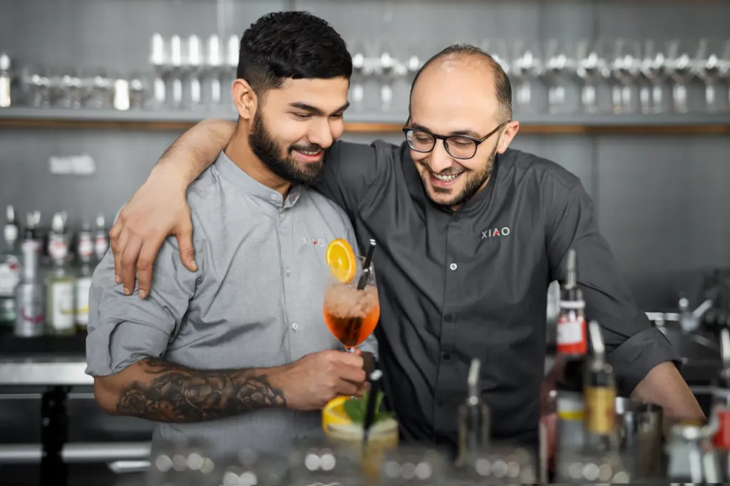 Ein junger Barman mit Tattoos hält einen Aperol-Spritz und hat Spaß mit seinem älteren Kollegen hinter der Theke