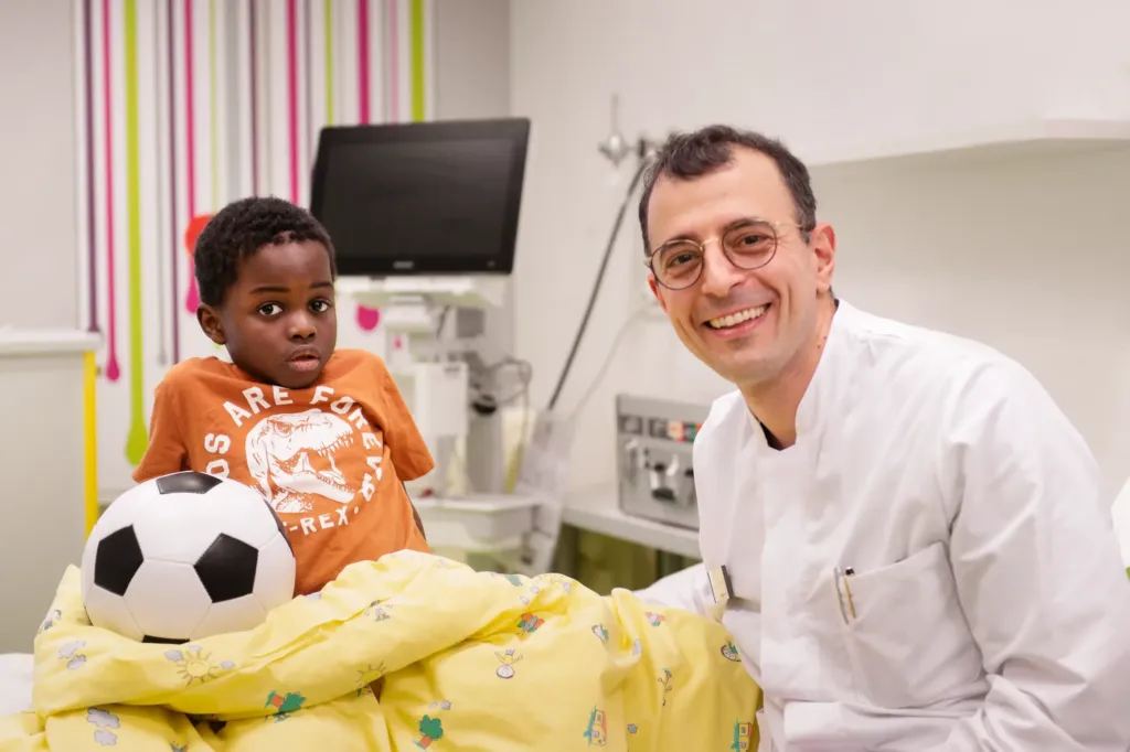 Ein Kind sitzt mit einem Fußball auf einem Krankenhausbett und ein Arzt lächelt in die Kamera.