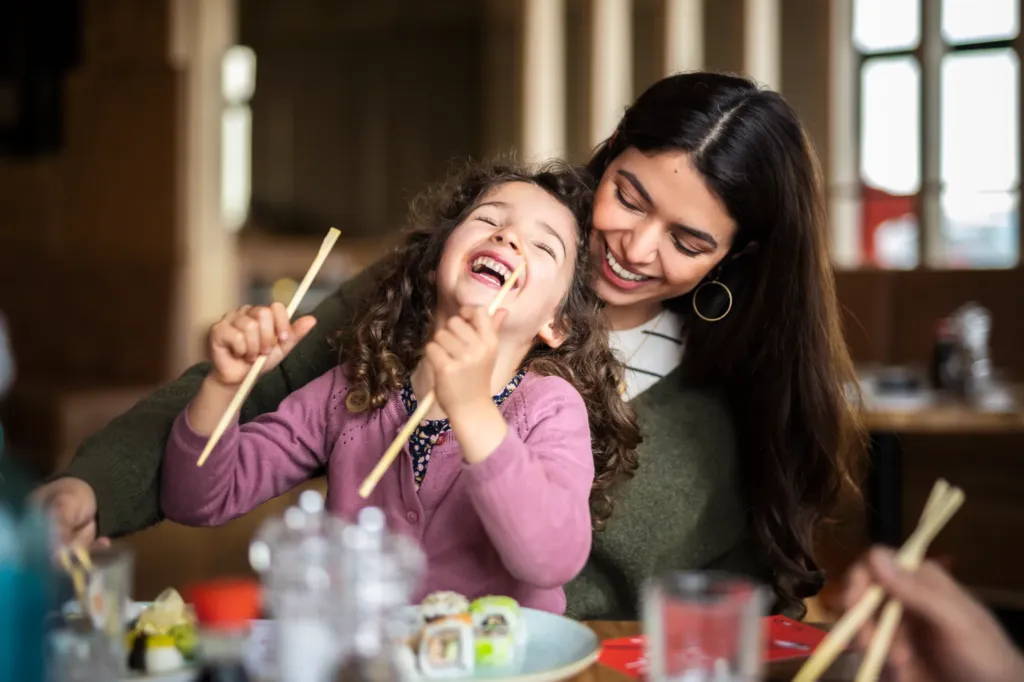 Ein lachendes Kind sitzt auf dem Schoß seiner Mutter und versucht Sushi mit Stäbchen zu essen