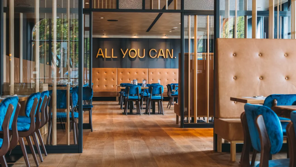 Gastraum des XIAO Hagen mit blauen Samtstühlen, gepolsterten Lederbänken und dem Schriftzug „All you can.“ an der Wand