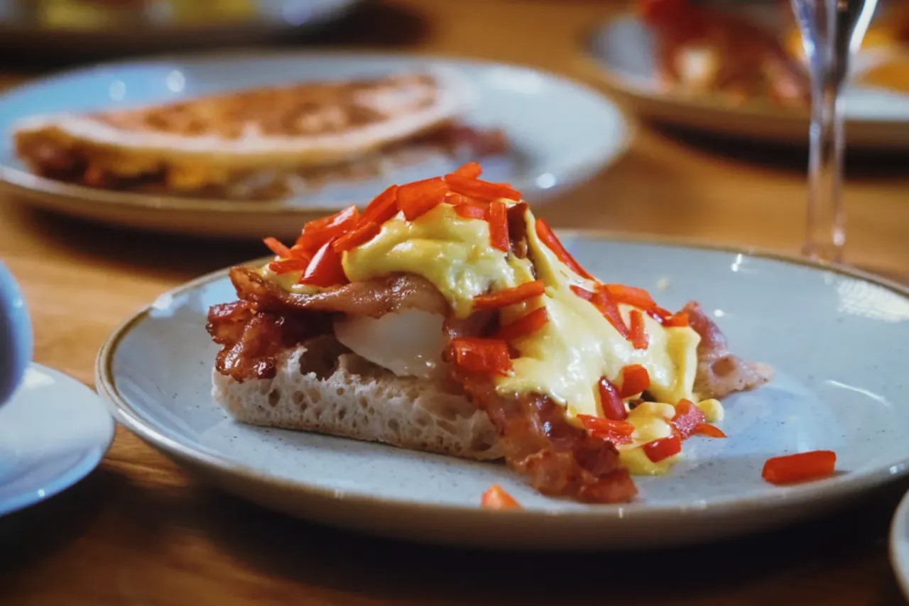 Brunch-Teller mit Eggs Benedict, Bacon und Sauce Hollandaise, im Hintergrund ein Glas Prosecco