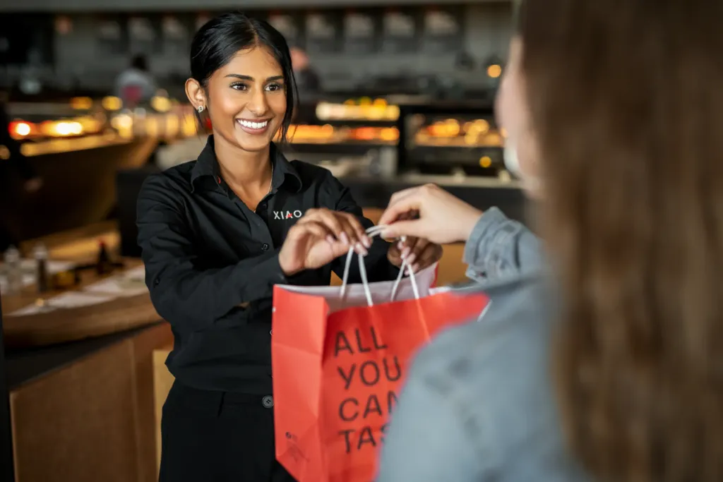 XIAO-Mitarbeiterin reicht einem Gast lächelnd eine rote Take-Away-Tüte über die Theke