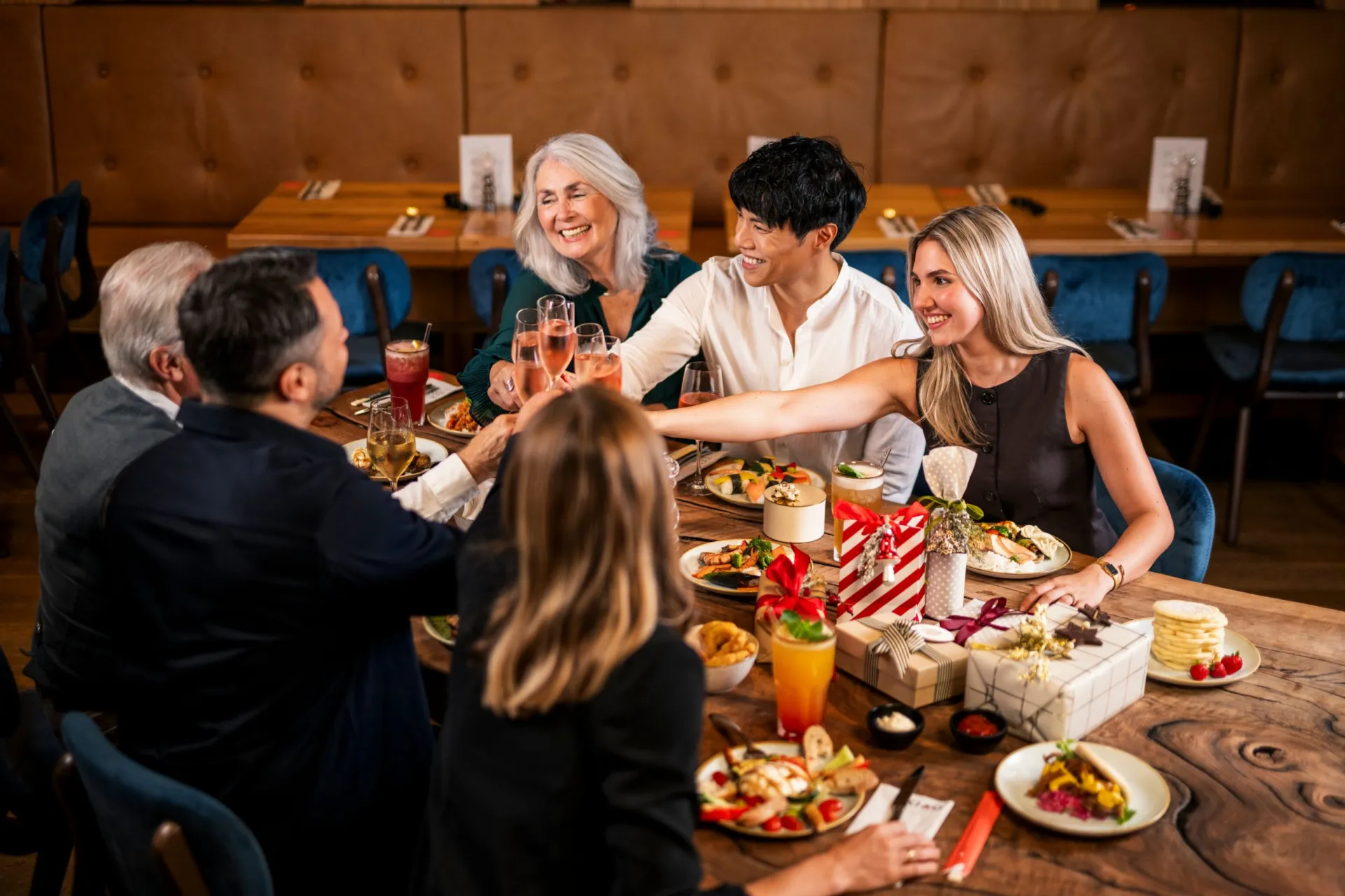 Sechs Gäste stoßen an einem Tisch im XIAO an – mit Tellern vom Buffet und weihnachtlich verpackten Geschenken